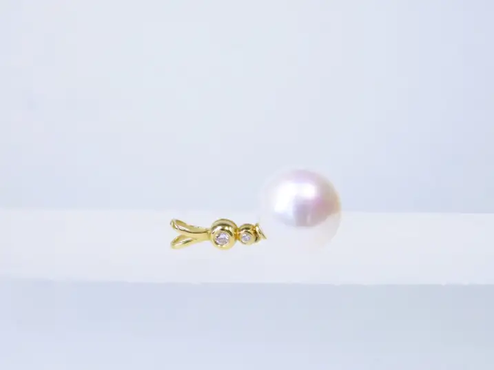 Diamantbesetzter Anhänger aus 14kt Gelbgold mit einer runden, weißen Mingperle in AAA-Qualität liegend auf einem Glasblock, Öse zeigt nach links.