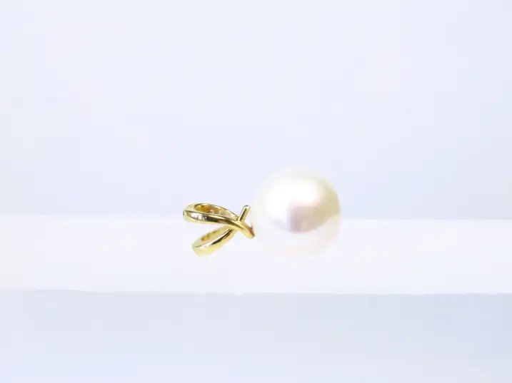 Geschwungener Anhänger aus 14kt Gelbgold mit einer runden, weißen Mingperle in AAA-Höchstqualität liegend auf einem Glasblock, Öse zeigt nach links.